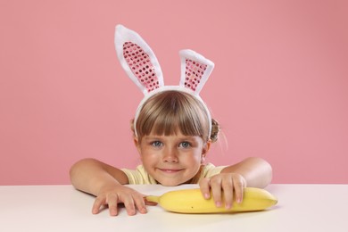 Photo of Cute little girl in bunny ears reaching for banana at white table against pink background