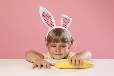 Photo of Cute little girl in bunny ears reaching for banana at white table against pink background