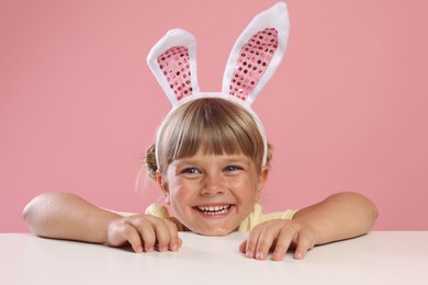 Photo of Curious little girl in bunny ears peeking over white table against pink background