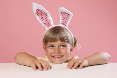 Photo of Curious little girl in bunny ears peeking over white table against pink background