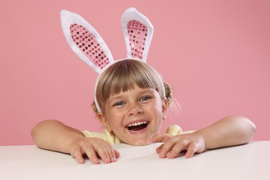 Photo of Curious little girl in bunny ears peeking over white table against pink background