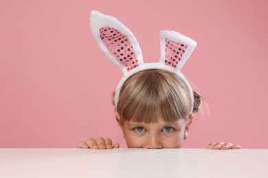 Photo of Curious little girl in bunny ears peeking over white table against pink background