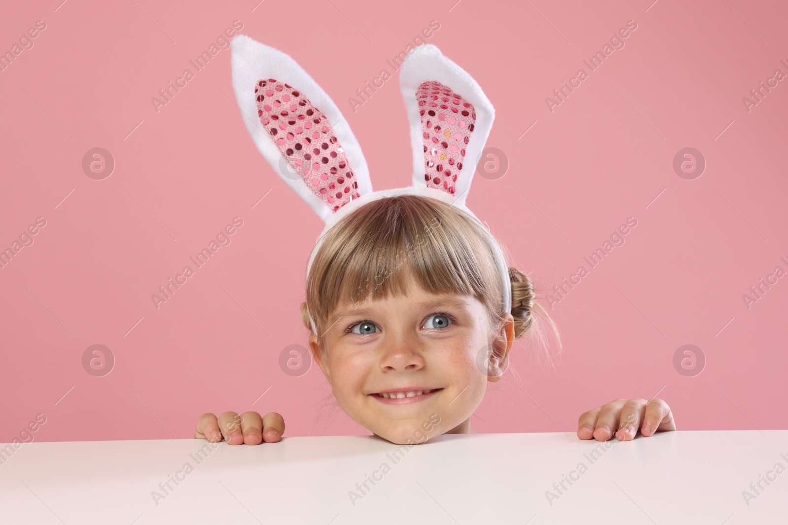 Photo of Curious little girl in bunny ears peeking over white table against pink background