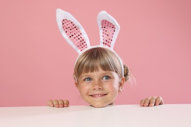 Photo of Curious little girl in bunny ears peeking over white table against pink background