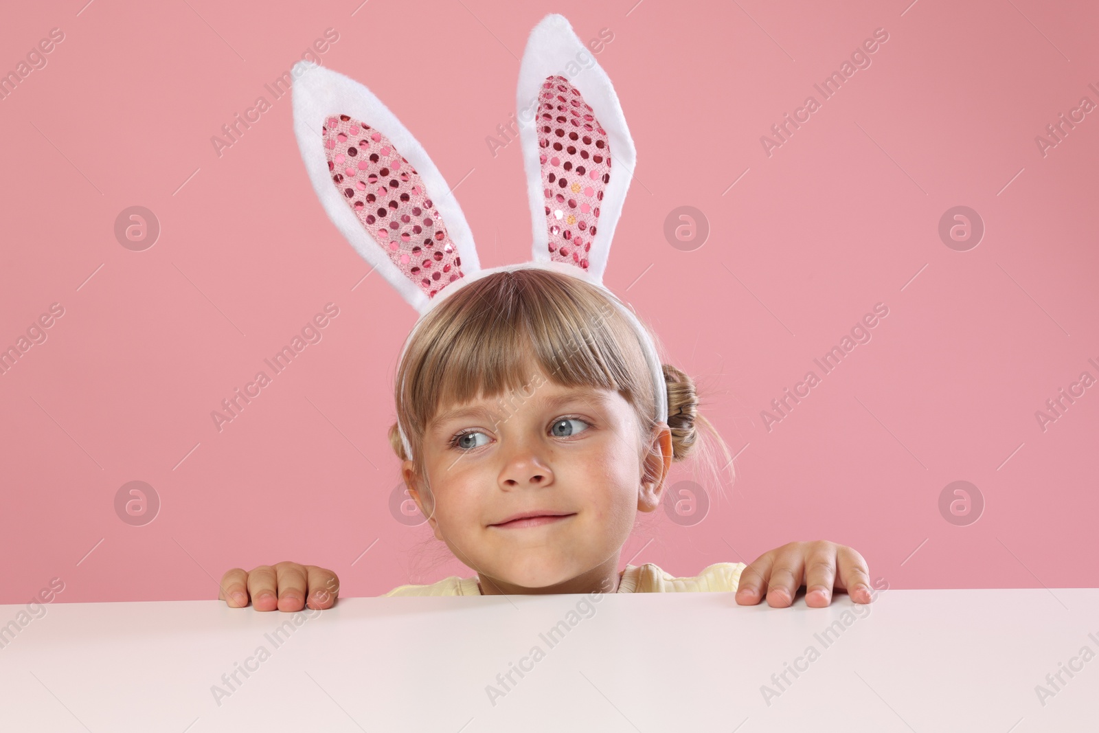 Photo of Curious little girl in bunny ears peeking over white table against pink background