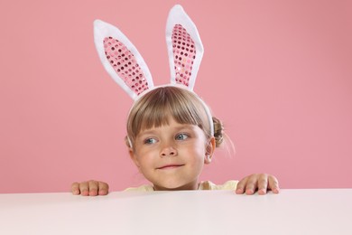 Photo of Curious little girl in bunny ears peeking over white table against pink background
