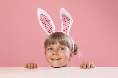 Photo of Curious little girl in bunny ears peeking over white table against pink background