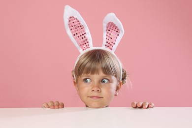 Photo of Curious little girl in bunny ears peeking over white table against pink background