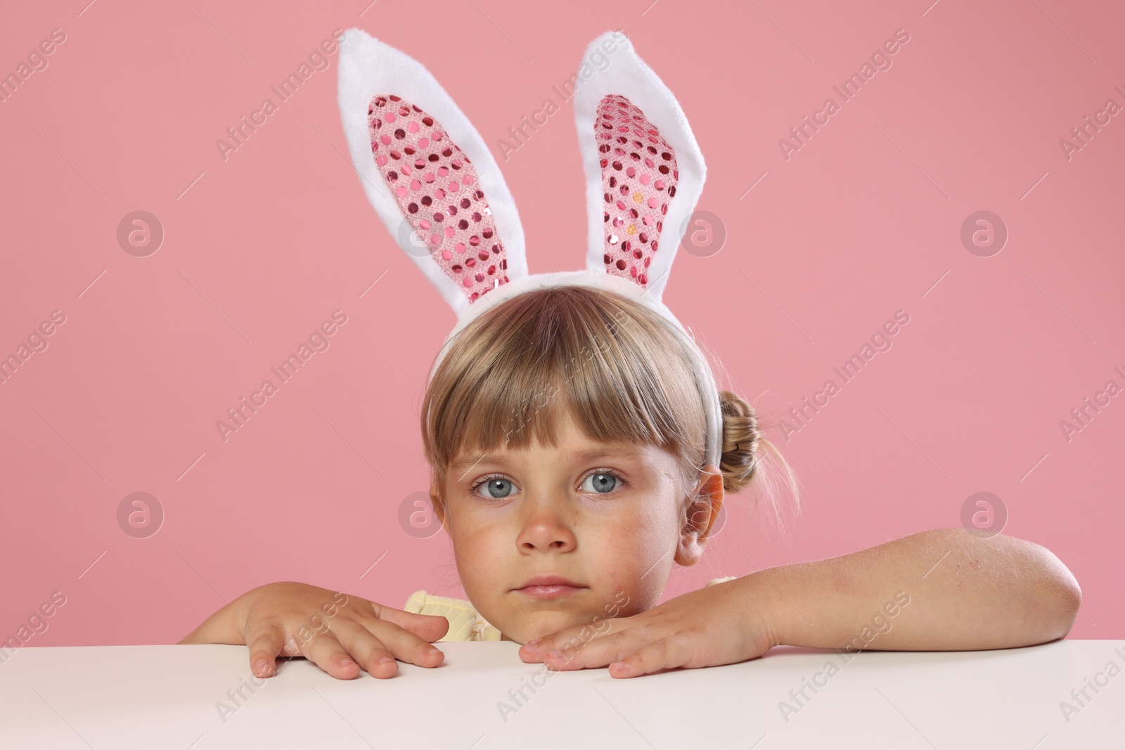 Photo of Curious little girl in bunny ears peeking over white table against pink background