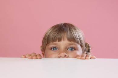 Photo of Curious little girl peeking over table against pink background