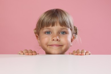 Photo of Curious little girl peeking over table against pink background