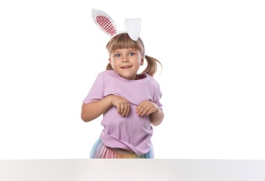 Photo of Curious little girl in bunny ears at table on white background