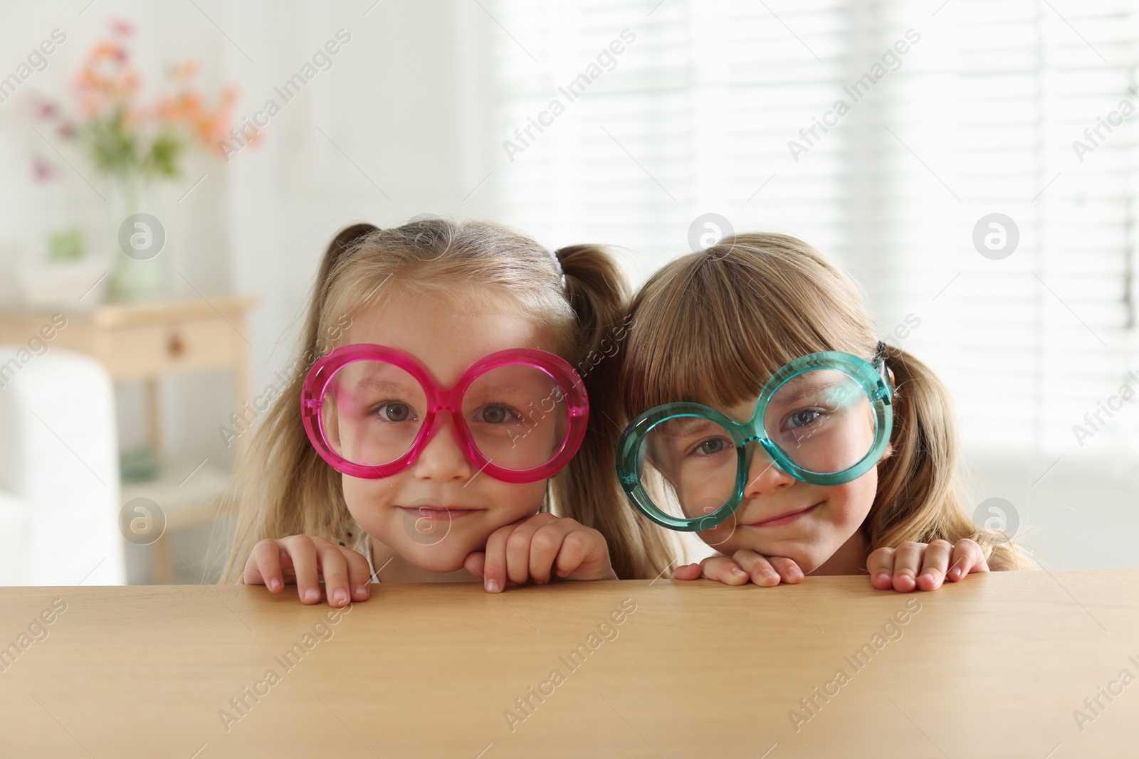 Photo of Curious little girls in funny glasses peeking over wooden table indoors