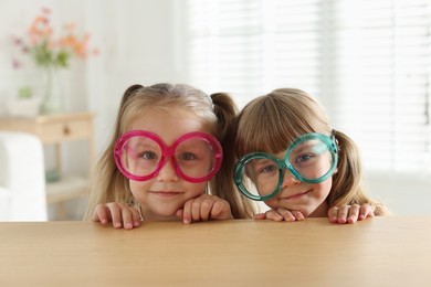 Photo of Curious little girls in funny glasses peeking over wooden table indoors