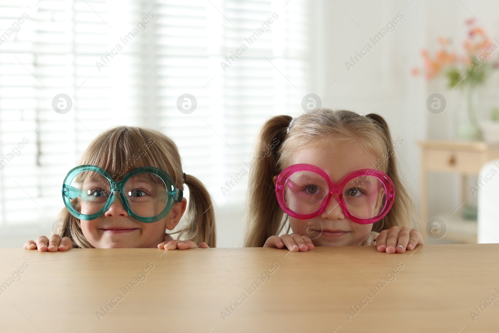 Photo of Curious little girls in funny glasses peeking over wooden table indoors