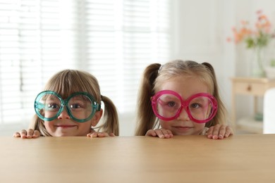 Photo of Curious little girls in funny glasses peeking over wooden table indoors