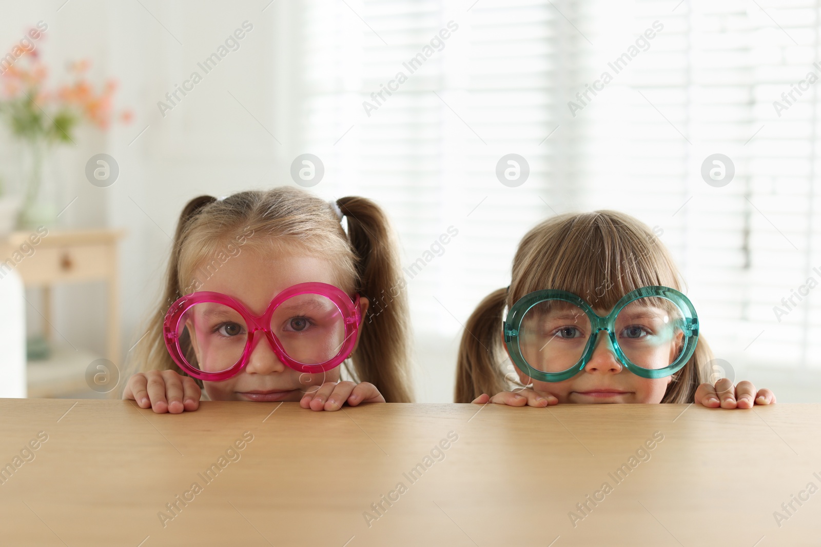 Photo of Curious little girls in funny glasses peeking over wooden table indoors
