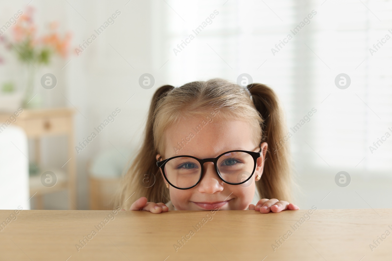 Photo of Curious little girl in glasses peeking over wooden table indoors