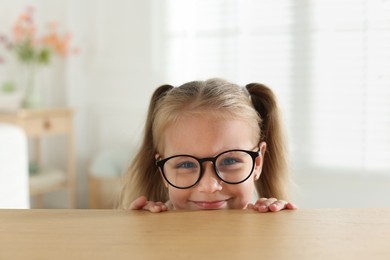 Photo of Curious little girl in glasses peeking over wooden table indoors