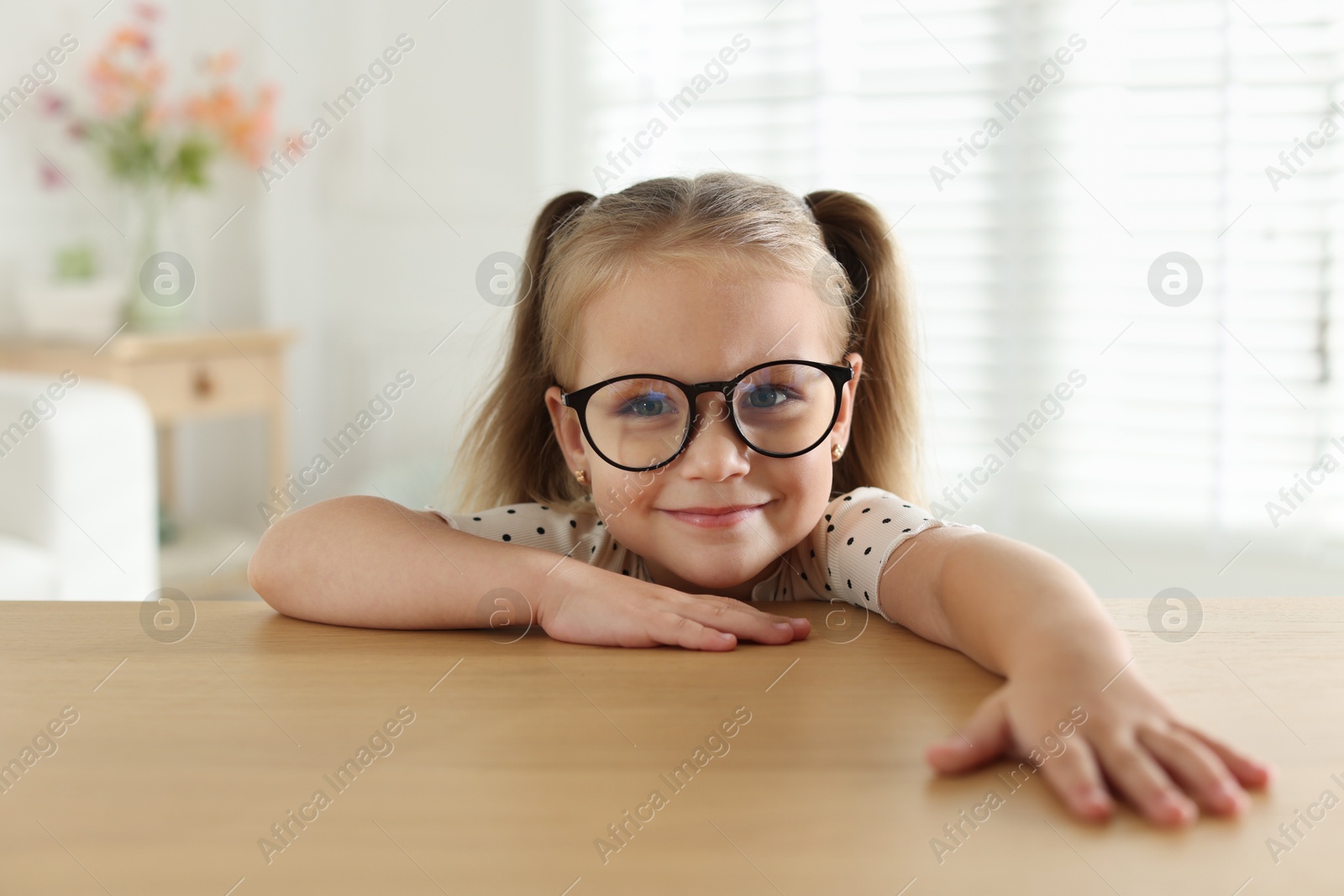 Photo of Curious little girl in glasses peeking over wooden table indoors