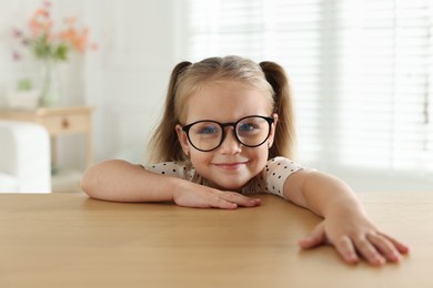 Photo of Curious little girl in glasses peeking over wooden table indoors