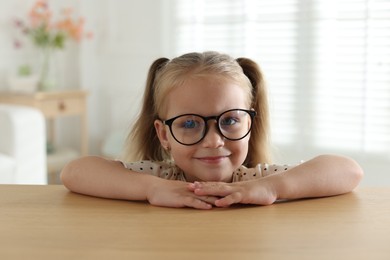 Photo of Curious little girl in glasses peeking over wooden table indoors
