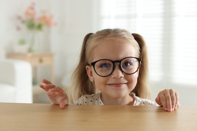 Photo of Curious little girl in glasses peeking over wooden table indoors