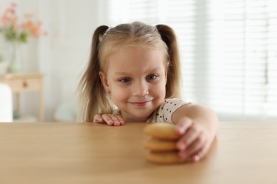 Photo of Cute little girl reaching for tasty cookies at wooden table indoors