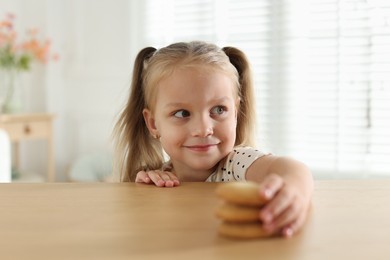 Photo of Cute little girl reaching for tasty cookies at wooden table indoors