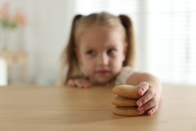 Photo of Cute little girl reaching for tasty cookies at wooden table indoors, selective focus