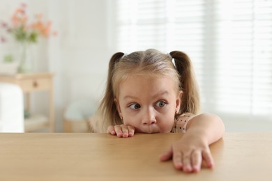 Photo of Curious little girl peeking over wooden table indoors, space for text