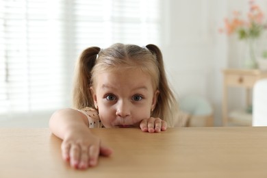Photo of Curious little girl peeking over wooden table indoors