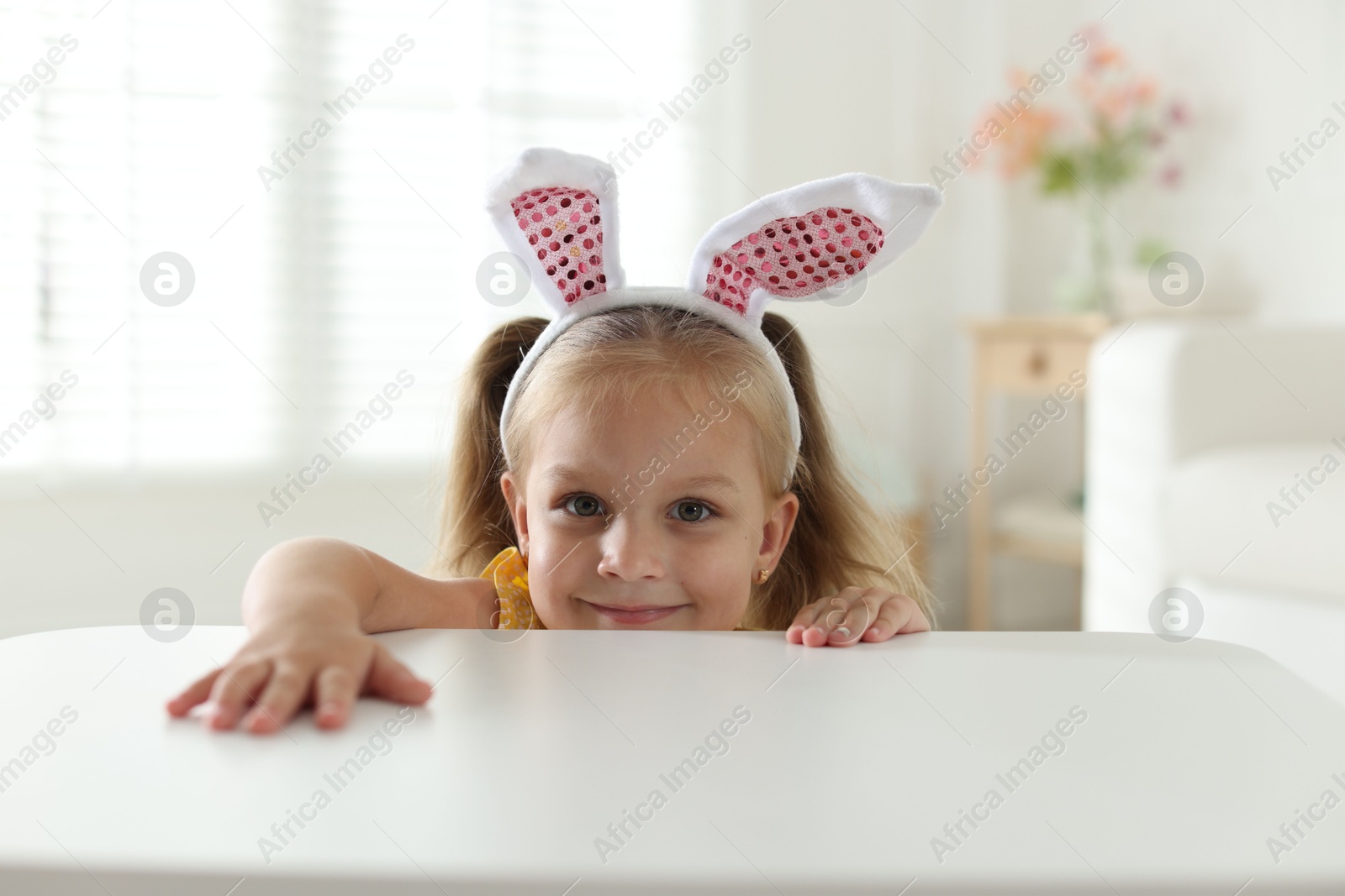 Curious little girl in bunny ears peeking over white table indoors Photo of Curious little girl in bunny ears peeking over white table indoors