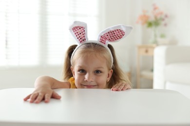 Photo of Curious little girl in bunny ears peeking over white table indoors