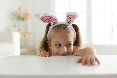 Photo of Curious little girl in bunny ears peeking over white table indoors
