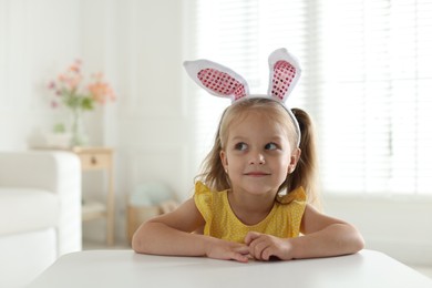 Photo of Curious little girl in bunny ears at white table indoors