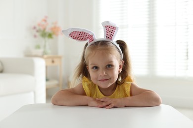 Photo of Curious little girl in bunny ears at white table indoors