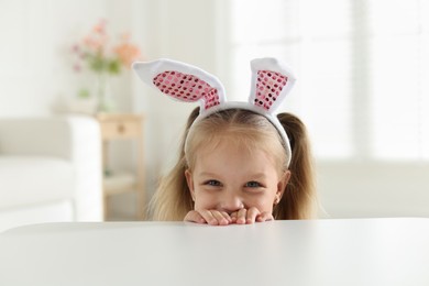 Photo of Curious little girl in bunny ears peeking over white table indoors
