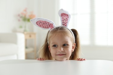 Photo of Curious little girl in bunny ears peeking over white table indoors