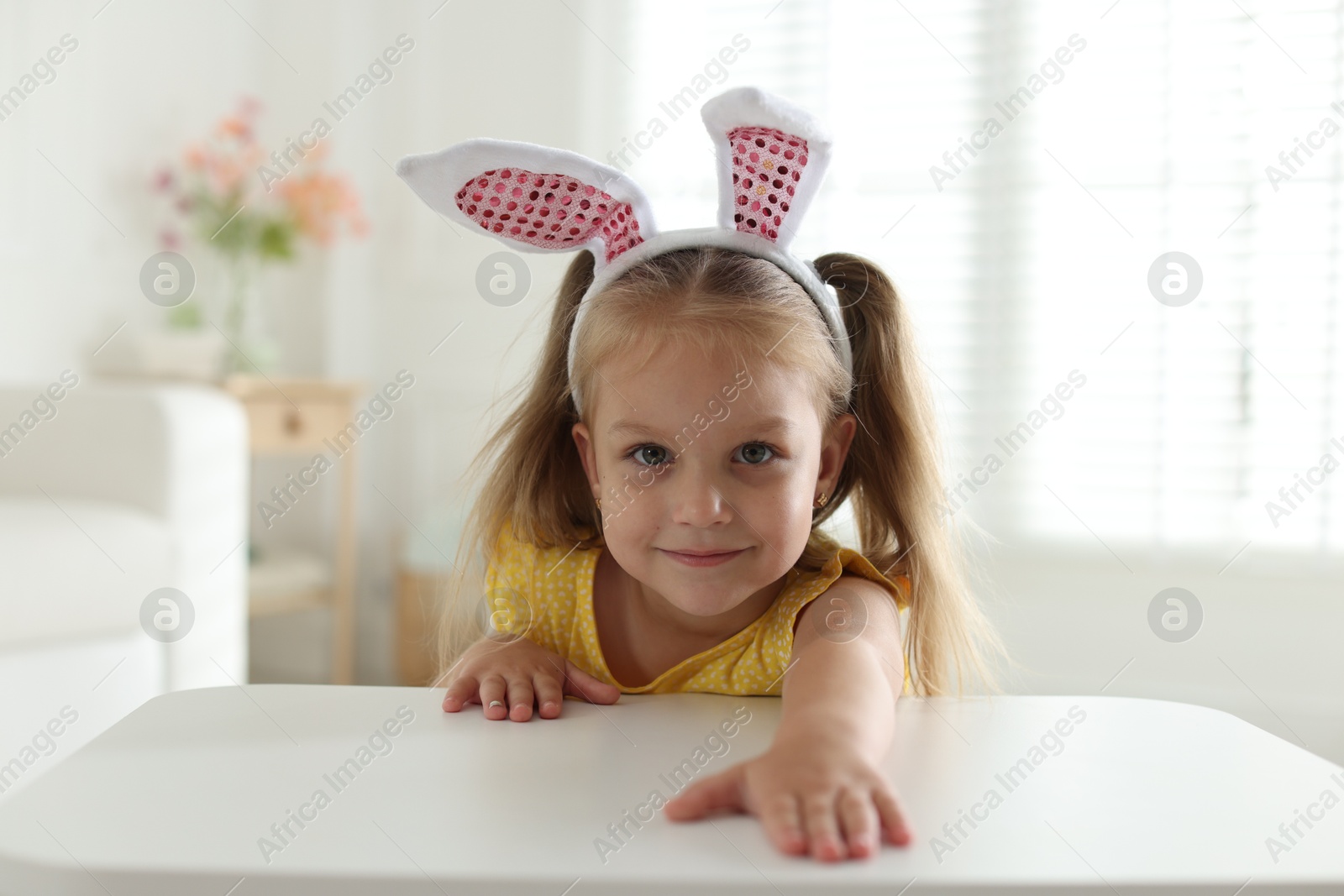 Curious little girl in bunny ears peeking over white table indoors Photo of Curious little girl in bunny ears peeking over white table indoors