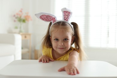 Photo of Curious little girl in bunny ears peeking over white table indoors