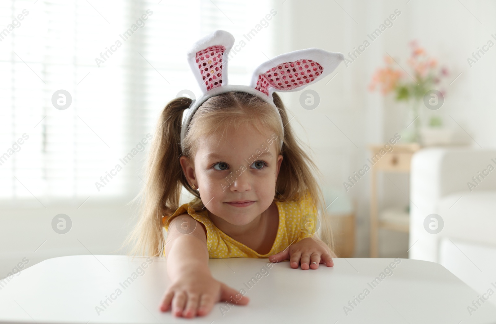 Photo of Curious little girl in bunny ears peeking over white table indoors