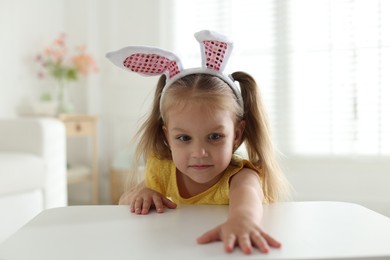 Photo of Curious little girl in bunny ears peeking over white table indoors