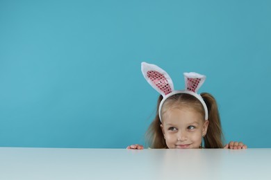 Photo of Curious little girl in bunny ears peeking over white table against light blue background, space for text
