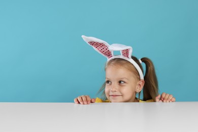 Photo of Curious little girl in bunny ears peeking over white table against light blue background, space for text