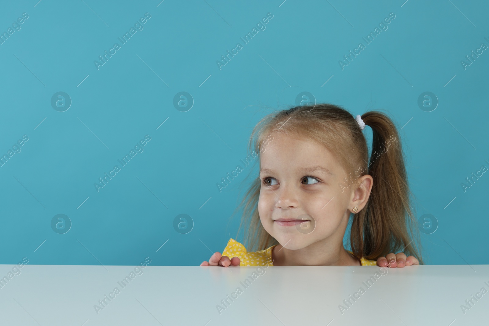 Curious little girl peeking over white table against light blue background, space for text Photo of Curious little girl peeking over white table against light blue background, space for text