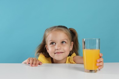 Photo of Cute little girl reaching for glass of orange juice at white table against light blue background