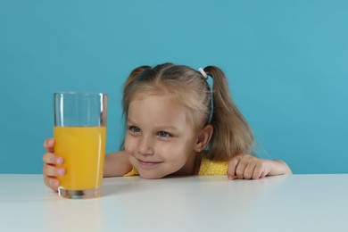 Photo of Cute little girl reaching for glass of orange juice at white table against light blue background