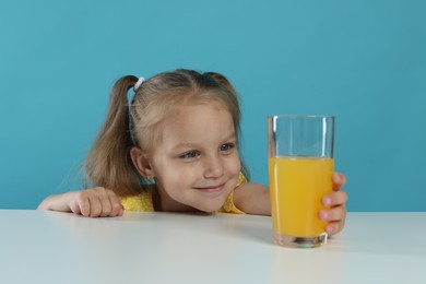 Photo of Cute little girl reaching for glass of orange juice at white table against light blue background