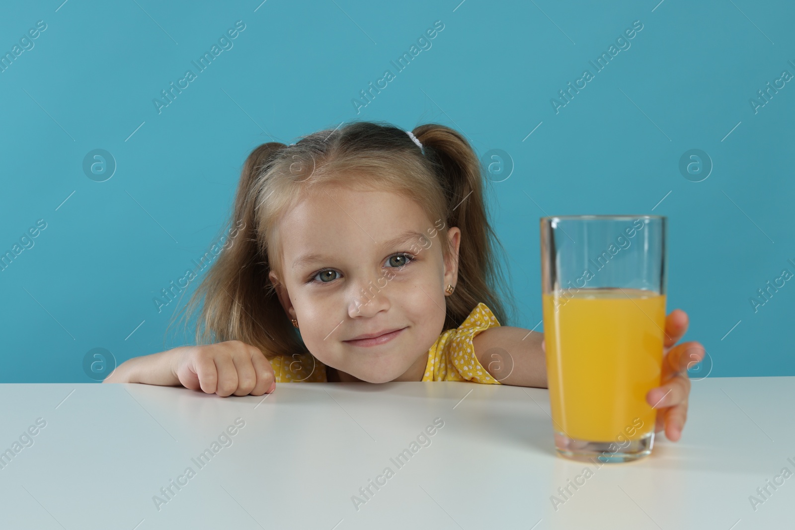 Photo of Cute little girl reaching for glass of orange juice at white table against light blue background
